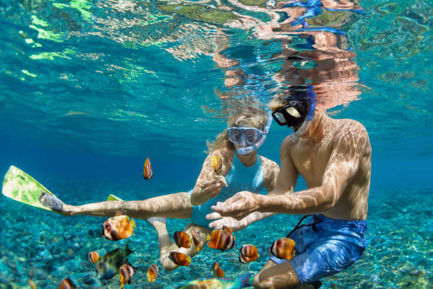 An underwater eye-level view of a man and a woman snorkeling in crystal-clear blue tropical water, surrounded by colorful fish and a sandy seabed beneath them