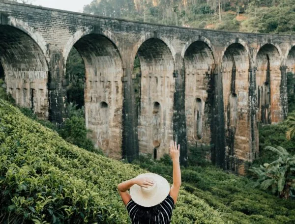 A tourist wearing a wide-brimmed hat stands in lush greenery, gazing at the iconic Nine Arches Bridge in Ella, Sri Lanka, surrounded by a scenic, forested landscape.